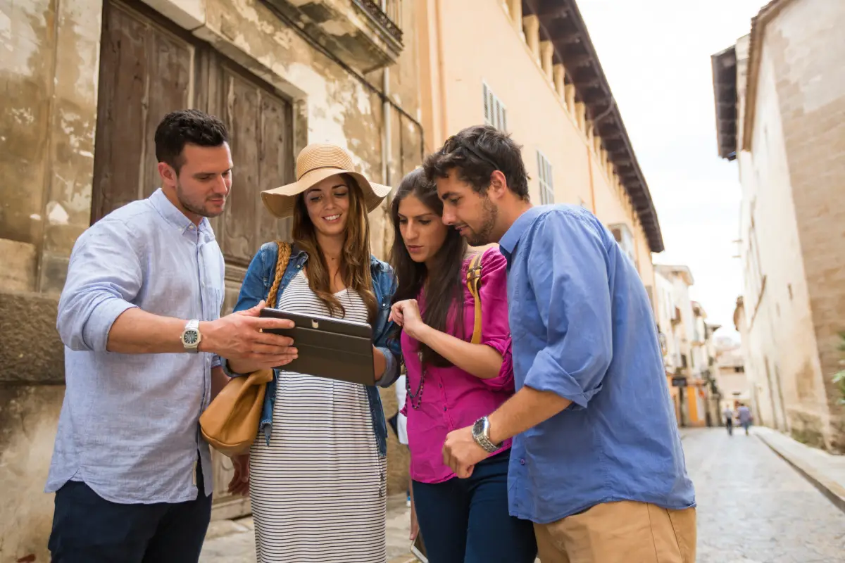 Cuatro amigos jóvenes consultando un mapa digital en una tablet mientras recorren una calle con edificios antiguos