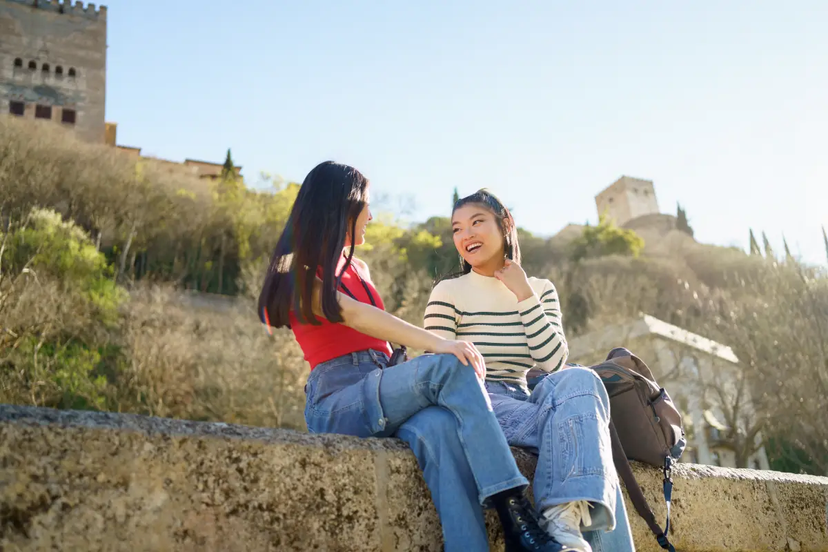 Dos jóvenes sentadas en un murete de piedra charlando alegremente, con una torre histórica y vegetación de fondo en un día soleado en Granada