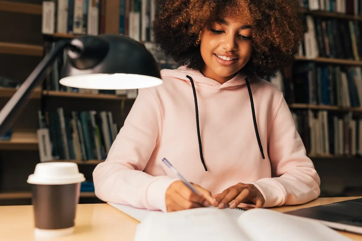 Estudiante tomando apuntes en una biblioteca con expresión concentrada y luz de sobremesa