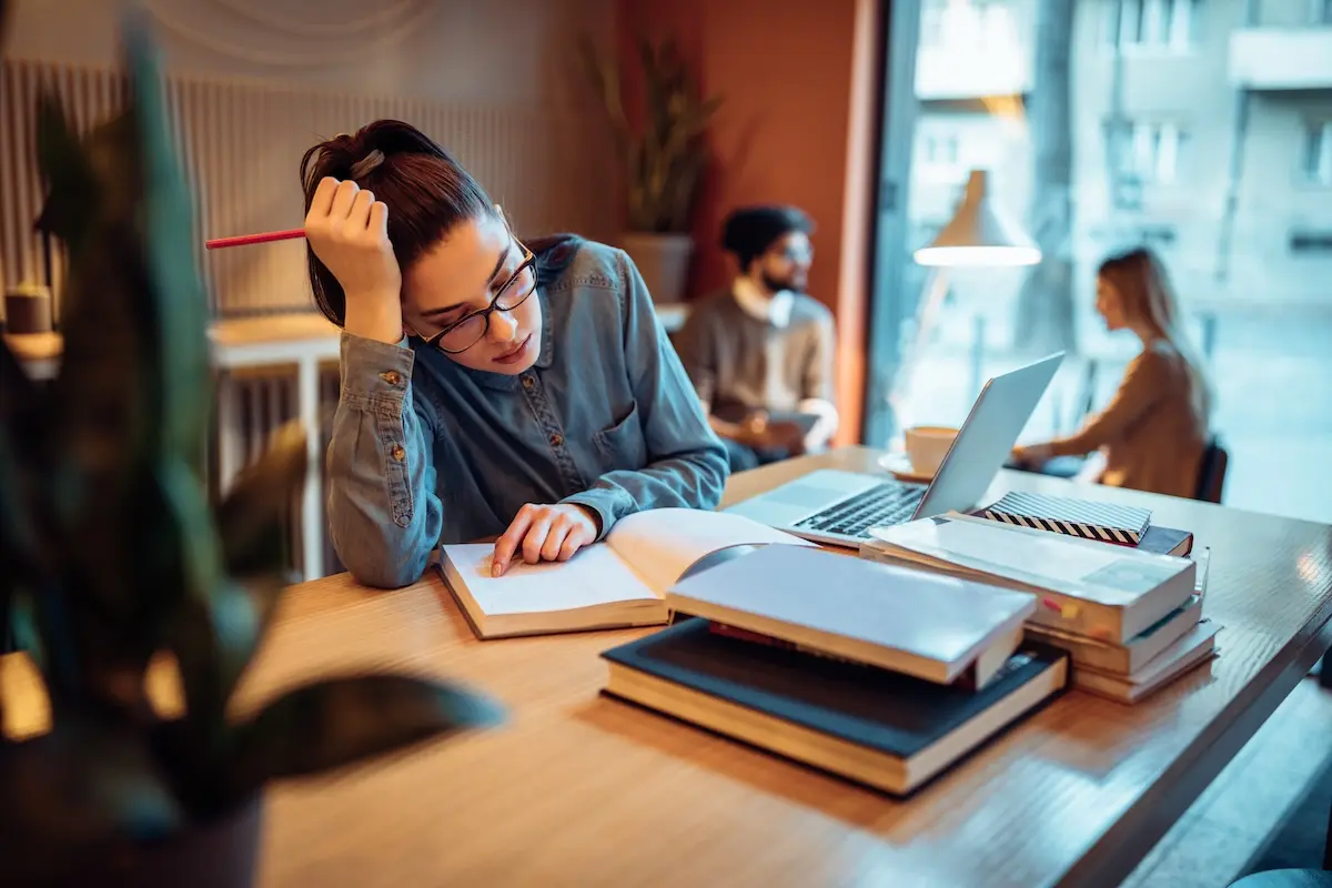 Estudiante cansada repasando un libro y apuntes en una mesa de estudio con portátil
