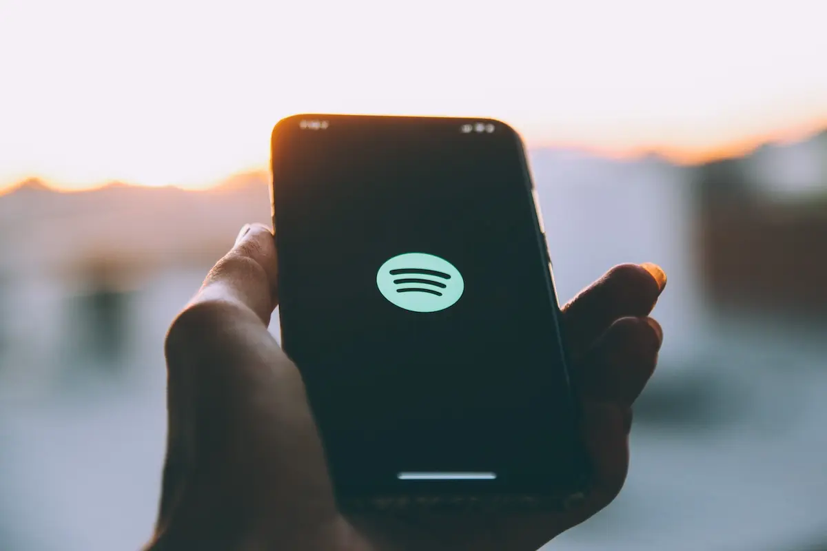 Mano sosteniendo un móvil con una aplicación de música abierta y auriculares sobre la mesa